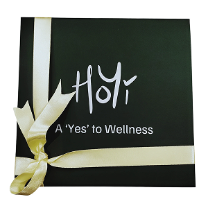 A black gift box with white text reading 'Holi' and 'A 'Yes' to Wellness', tied with a yellow ribbon. The box contains a custom pack of 3 preserves and crochet toys.