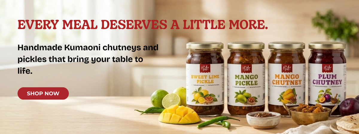 Four jars of RAK Handmade Kumaoni chutneys and pickles displayed on a wooden table with fresh ingredients like mango, lime, and onions. The background is a warm, softly blurred kitchen setting with red promotional text reading 'EVERY MEAL DESERVES A LITTLE MORE.