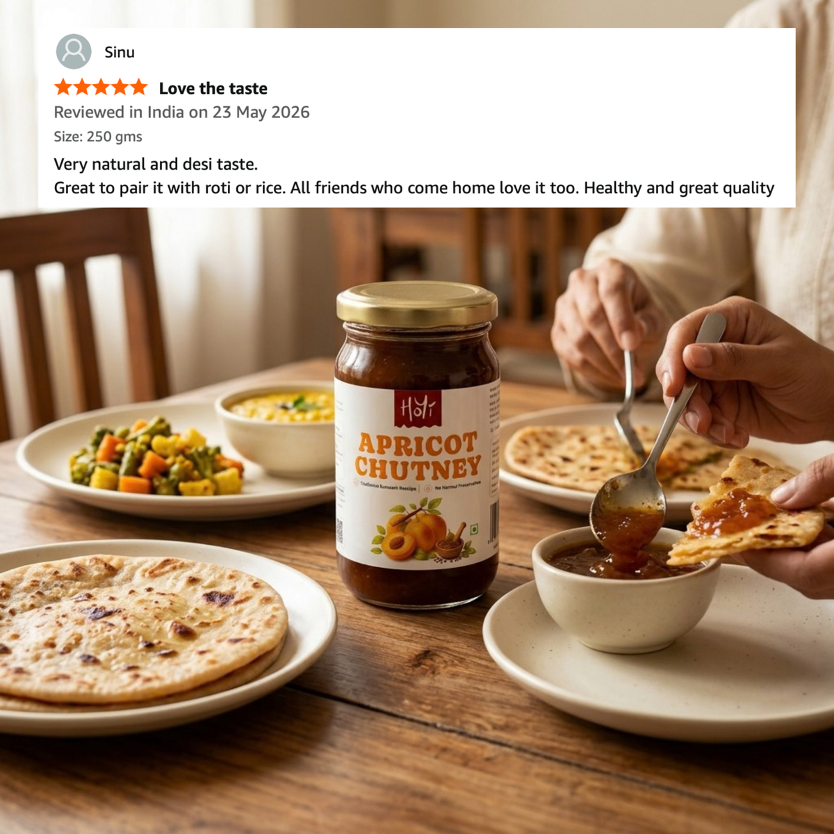 A jar of apricot chutney sits on a wooden dining table, surrounded by flatbreads, a bowl of chutney, and a plate of mixed vegetables. A person is visible on the right, holding a spoon and spreading the chutney onto a piece of bread.